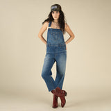 A woman models The Juniper Overall by Sendero Provisions Co.—featuring a raw hem—paired with a trucker hat and red cowboy boots, standing against a plain beige background with one leg crossed over the other.