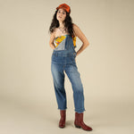 A woman wearing The Juniper Overall by Sendero Provisions Co.—styled with one strap undone and a raw hem—paired with a red cap, yellow printed top, and red cowboy boots, stands against a neutral background.