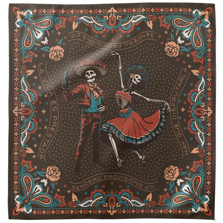 Introducing the Viejones Bandana by Sendero Provisions Co.—a square bandana adorned with stylized skeletons inspired by Los Viejones and the traditional Tejas skeleton dance. Each figure, dressed in festive attire to honor ancestral celebrations, is surrounded by vibrant floral patterns and Spanish text set against a dark background.