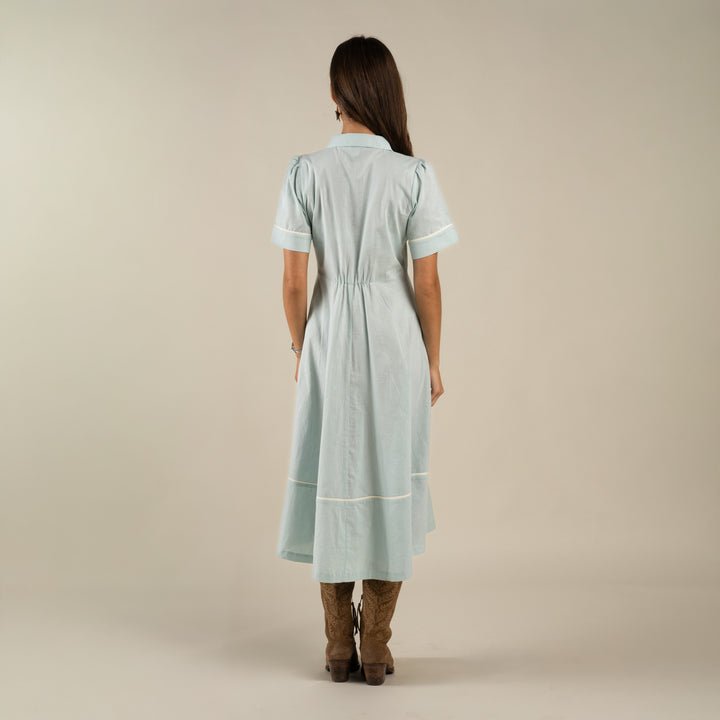 A woman stands facing away, wearing the light blue, short-sleeved La Dahlia Dress by Sendero Provisions Co.—crafted from hemp cotton with white trim—paired with brown knee-high boots, set against a plain beige background.