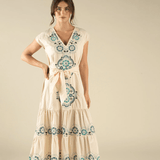 A woman stands against a beige background wearing The Emilia Dress by Sendero Provisions Co.—a flowy cotton dress with blue and green embroidery, waist tie, and tiered fit.