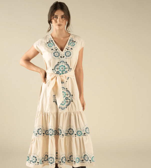 A woman stands against a beige background wearing The Emilia Dress by Sendero Provisions Co.—a flowy cotton dress with blue and green embroidery, waist tie, and tiered fit.