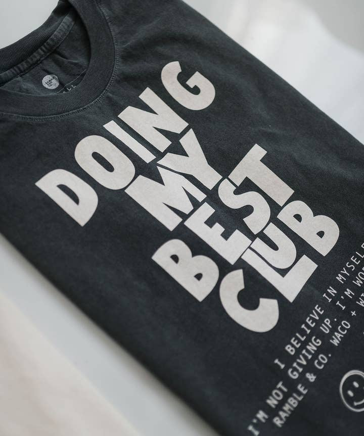 Black vintage faded t-shirt from Ramble & Company featuring bold white “DOING MY BEST CLUB” text, smaller lettering at the bottom, and a smiley face graphic. The Doing My Best Club tee offers a boxy, relaxed silhouette for effortless laid-back style.