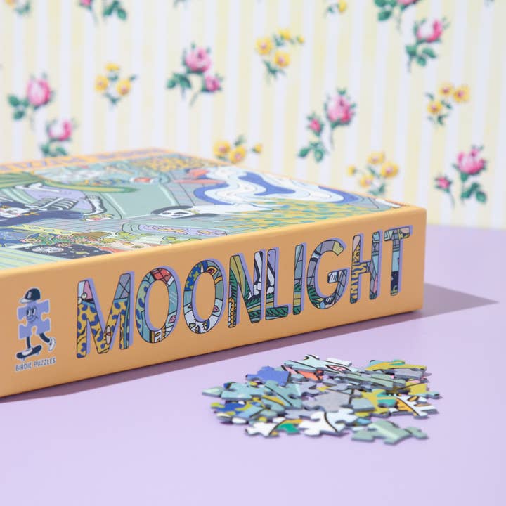 A box labeled "Moonlight 1,000 Piece Puzzle | Birdie Puzzles" sits on a lavender surface beside artist-designed puzzle pieces, with floral-patterned wallpaper in the background.