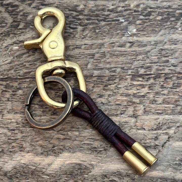 The Flint Trigger Keychain by Tres Cuervos features a brass clip with a key ring and two leather straps joined together, each finished with brass caps that accentuate the rich leather patina, and is completed with a handcrafted tassel—all presented on a wooden surface.