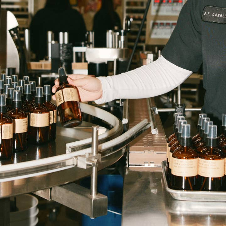 A person arranges amber bottles of N0.32 Sandalwood Rose Room & Linen Spray by P.F Candle, with beige labels, on a conveyor belt in a production facility.
