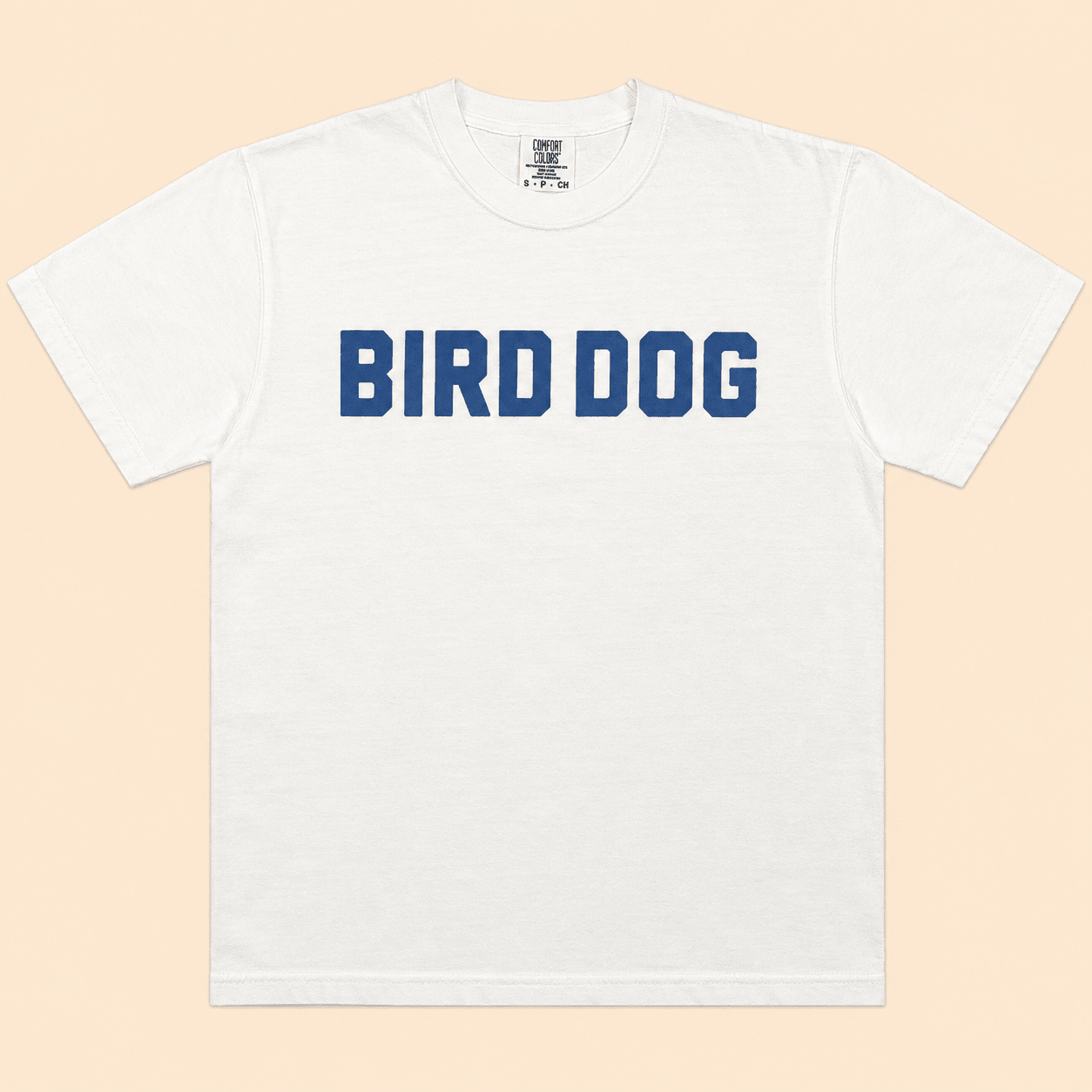Bird Dog Honor Shirt by Ballad Originals—crafted from premium USA cotton, this white shirt features “BIRD DOG” printed in bold blue letters across the chest and is displayed flat on a light beige background. A must-have for enthusiasts of classic hunting apparel.
