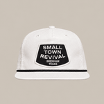 The Small Town Revival Patch Hat by Ballad Originals is a white baseball cap featuring a black patch on the front that reads “SMALL TOWN REVIVAL BRENHAM TEXAS.” Displayed against a plain beige background, this classic snapback design is perfect for showing your Brenham pride.