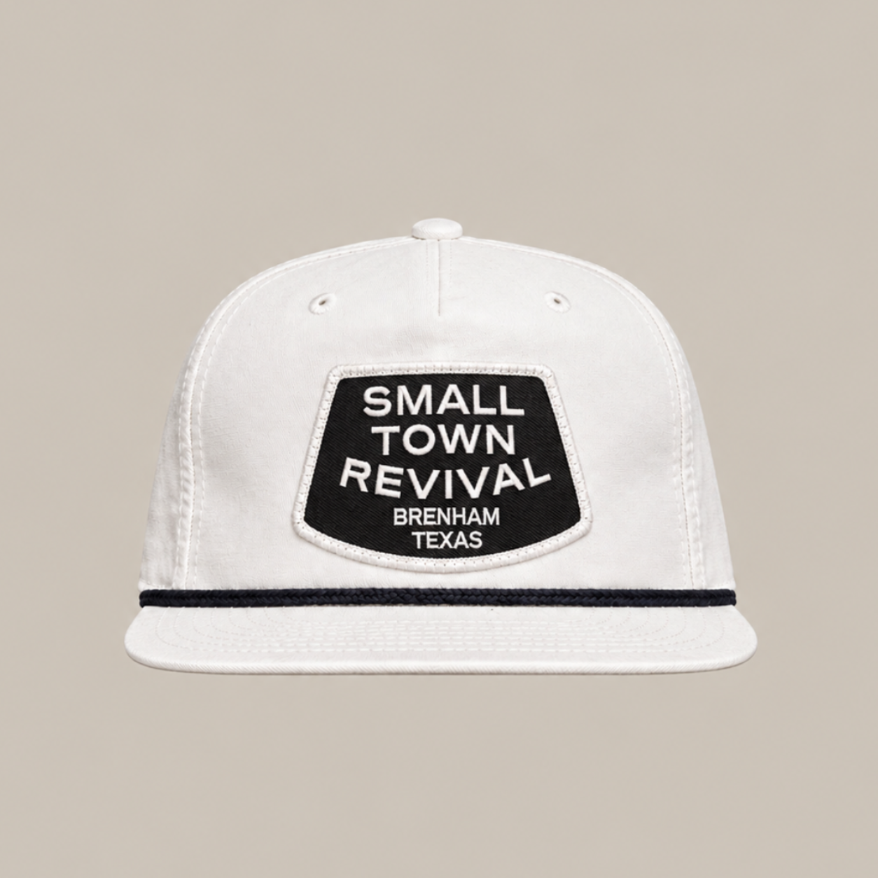 The Small Town Revival Patch Hat by Ballad Originals is a white baseball cap featuring a black patch on the front that reads “SMALL TOWN REVIVAL BRENHAM TEXAS.” Displayed against a plain beige background, this classic snapback design is perfect for showing your Brenham pride.