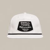 The Small Town Revival Patch Hat by Ballad Originals is a white baseball cap featuring a black patch on the front that reads “SMALL TOWN REVIVAL BRENHAM TEXAS.” Displayed against a plain beige background, this classic snapback design is perfect for showing your Brenham pride.