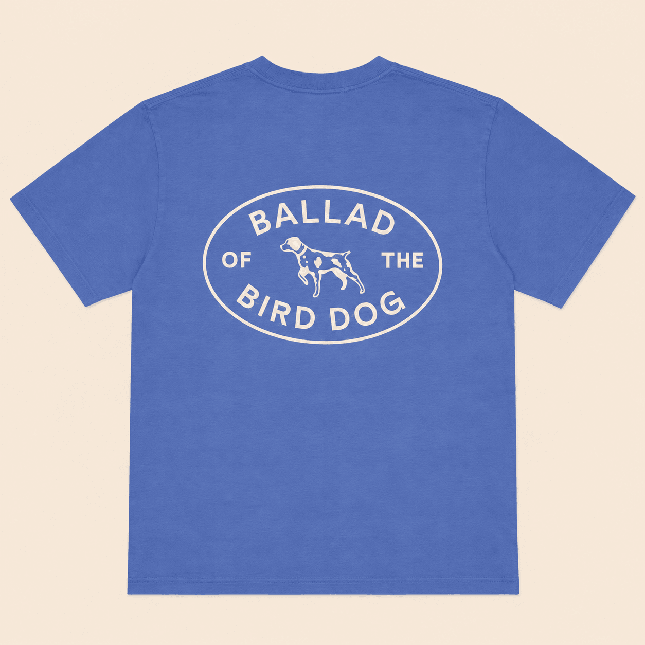 The Bird Dog Emblem Shirt by Ballad Originals features a blue design with an oval graphic on the back that reads “Ballad of the Bird Dog” and showcases a simple dog illustration at the center—perfect for displaying your bird dog pride while supporting local business.