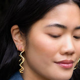 A woman with long dark hair looks downward calmly, wearing the Alma Hoops – 18K Gold Plated by Peter and June, along with a small stud earring.