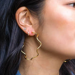Close-up of a woman's ear with three earrings: a small stud, a thin hoop, and the Alma Hoops - 18K Gold Plated by Peter and June—a large, wavy waterproof hoop.