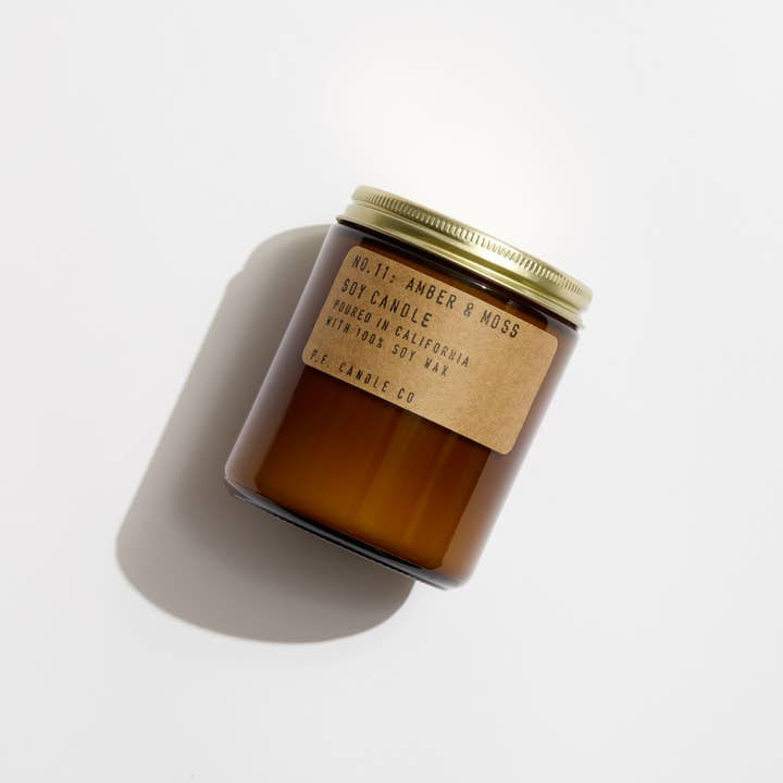 Amber glass jar with a gold lid and brown label reading "N0.11 Amber & Moss | P.F Candle" by P.F Candle, hand-poured soy candle made with 100% soy wax, shown on a white background.