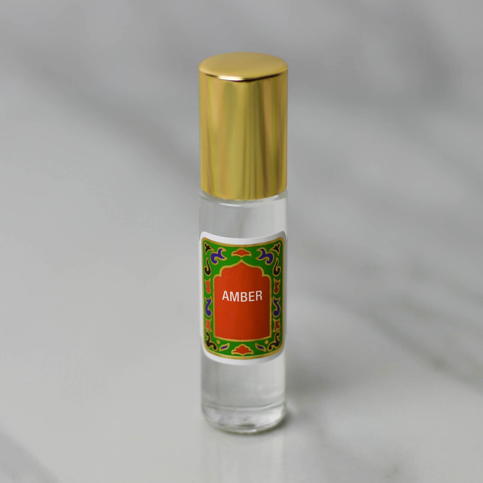 Amber Perfume Oil by Nemat is a small glass bottle with a gold cap and a decorative "Amber" label, resting on light gray marble. This subtle scent is perfect as your personal fragrance.