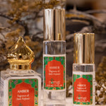 Three bottles of Nemat Amber Perfume Oil, each with gold and silver caps, are displayed on a surface. The subtle personal scent is highlighted, with dried flowers softly blurred in the background.