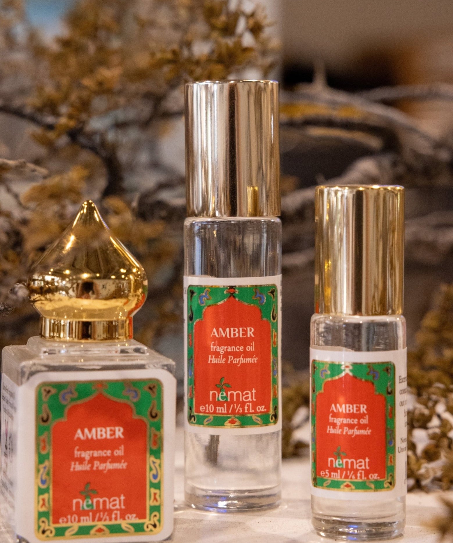 Three bottles of Nemat Amber Perfume Oil, featuring red and green labels, are displayed in front of dried flowers and a soft-focus background—inviting you to discover your unique scent with this subtle fragrance by Nemat.