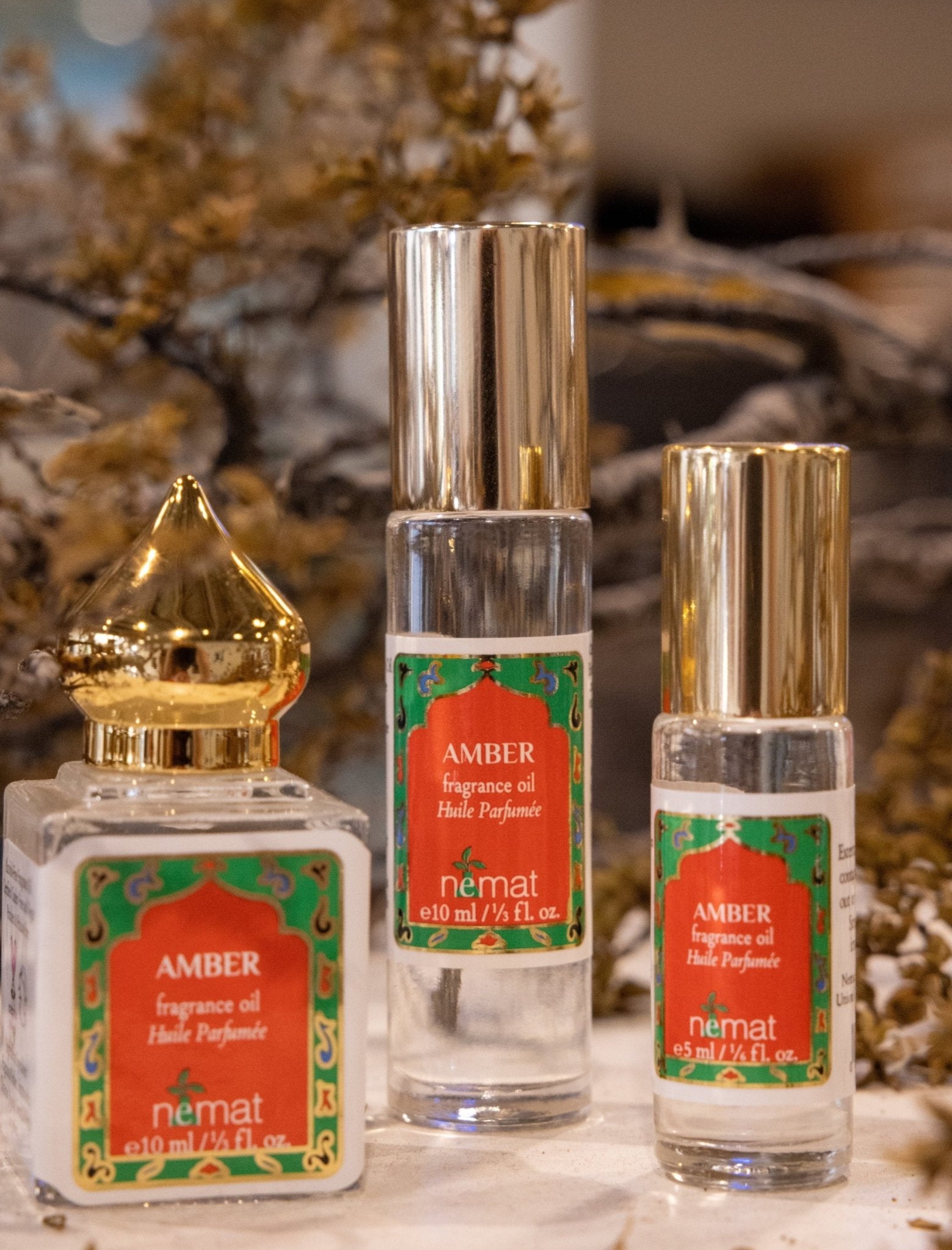 Three bottles of Nemat Amber Perfume Oil, each with gold and silver caps, are displayed on a surface. The subtle personal scent is highlighted, with dried flowers softly blurred in the background.