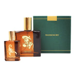 Two amber glass perfume bottles with gold caps sit before a green "Preservation West" box. American Ash by Preservation West, featuring a scorpion and a cowboy, evokes mysterious notes of Patchouli and Oud.