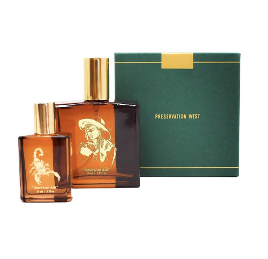 Two amber glass perfume bottles with gold caps—one featuring a scorpion, the other a cowboy—are displayed beside a green box labeled “American Ash” by Preservation West.