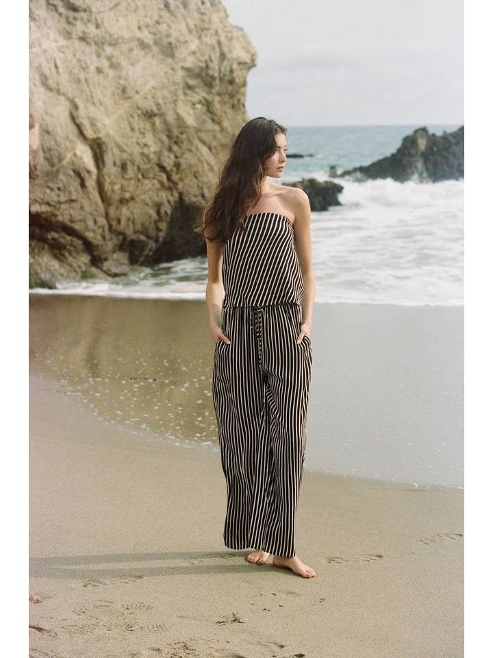 A woman wearing the Amina Pants by A. Ren stands barefoot on a sandy beach near the ocean, with rocky cliffs in the background.