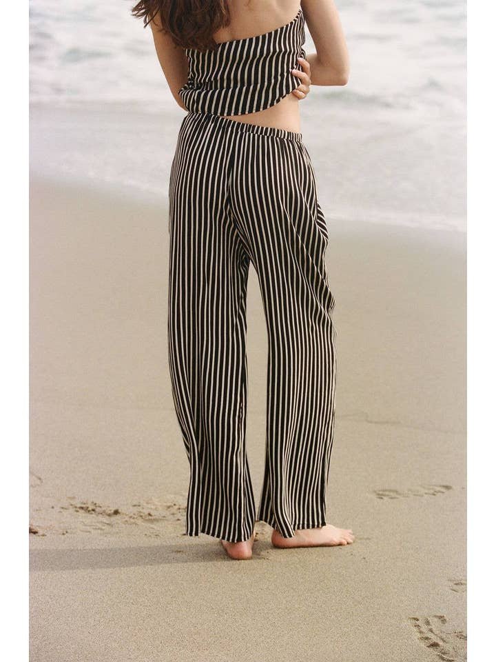 A person stands barefoot on the beach, gazing at the ocean while wearing A. Ren's Amina Pants—black and white striped wide leg pants paired with a matching top.