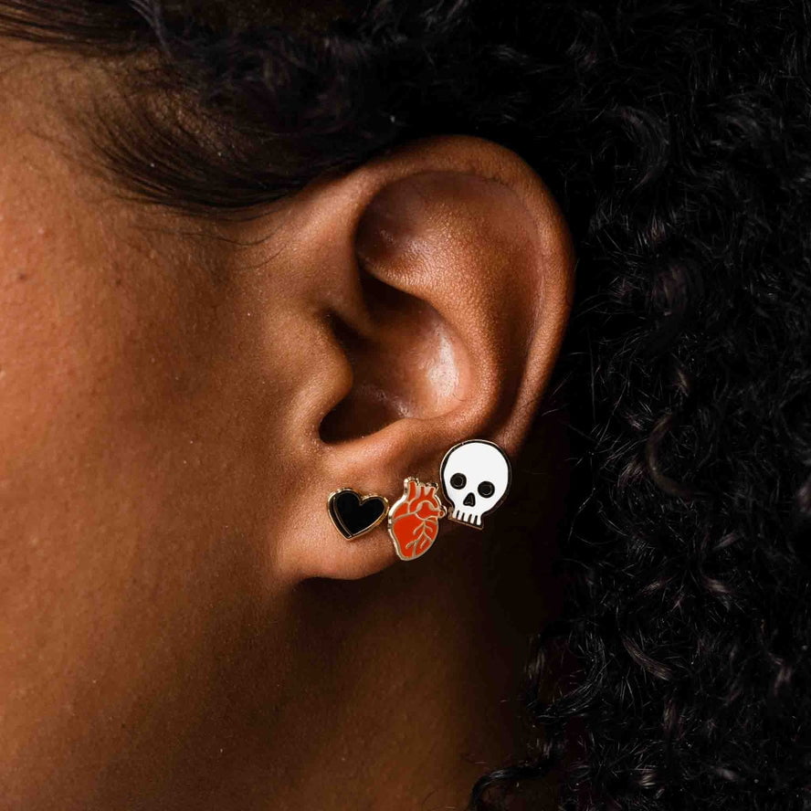 Close-up of an ear wearing three Larissa Loden studs: a black heart enamel stud, a red Anatomical Heart Stud from the Anatomical Heart Studs collection, and a white skull 18k gold-plated stud.