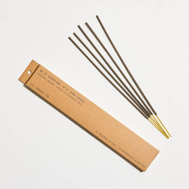 A brown box labeled “Arroyo Oak Incense Sticks | P.F Candle Co.” sits beside five brown and gold hand-dipped incense sticks, representing the signature California oak scent by P.F Candle.