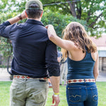 A man and woman stand on grass, both facing away. The woman has her arm around the man's shoulders, who wears a hand-stitched Argentine leather Zilker Belts ATX Dark belt. Trees and a building are seen in the background.