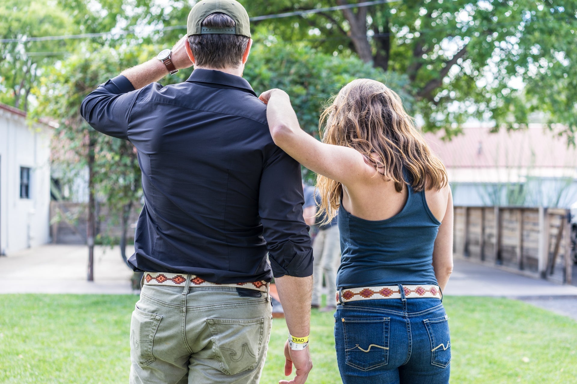 A man and woman stand on grass, both facing away. The woman has her arm around the man's shoulders, who wears a hand-stitched Argentine leather Zilker Belts ATX Dark belt. Trees and a building are seen in the background.