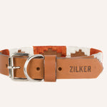 The ATX Dog Collar by Zilker Belts features brown leather, a white and orange patterned fabric band, a metal buckle, hand stitching, and “ZILKER” engraved for a stylish and durable finish.