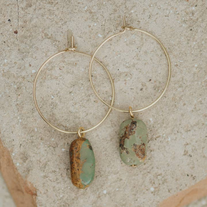 The Aurora Hoops by CommonForm feature brass hoops with oval, greenish-brown stone pendants, displayed on a textured beige surface—a stylish example of handmade jewelry.