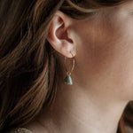 A close-up of a woman's ear wearing CommonForm's Aurora Hoops: handmade brass earrings featuring a green stone pendant.