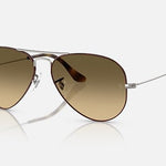 The Ray-Ban 3025 Aviator sunglasses feature a classic pilot shape with thin metal frames, curved arms, and the iconic logo on the lens, shown in brown tint against a white background.