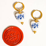 Yun Ceramic Floral Heart Earring by Flanerie by MAIQ: two hypoallergenic hoop earrings with white ceramic heart charms painted with blue floral patterns, next to a red quarter-sized coin.