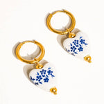 A pair of Yun Ceramic Floral Heart Earring by Flanerie by MAIQ, featuring 18K gold hoop earrings with white heart-shaped ceramic charms, blue floral patterns, and gold beads.