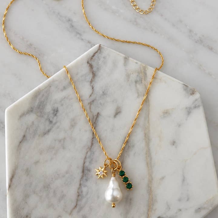 Introducing the Pearl, Cz Emerald, Star Charm Necklace by Mesa Blue—a refined 18K gold-plated chain showcasing a freshwater pearl pendant, a gold star charm, and three sparkling CZ emerald gemstones. Beautifully presented on a marble hexagon slab, this elegant necklace from Mesa Blue effortlessly elevates any look with its sophisticated design.