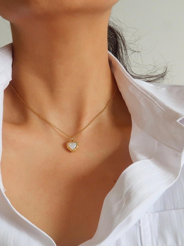 A person wearing the Josie Vintage Heart Pendant Necklace by Flanerie by MAIQ, a vintage heart pendant on a thin gold chain featuring a white center on the charm.