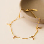 The Anklet | Champagne Drop by JaxKelly, featuring small baguette CZ stones on a delicate gold chain, rests near the rim of a beige ceramic dish on a light surface.