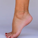 A close-up of a foot with red toenails wearing the JaxKelly Champagne Drop Anklet, featuring delicate baguette CZ stones and small charms, set against a plain background.