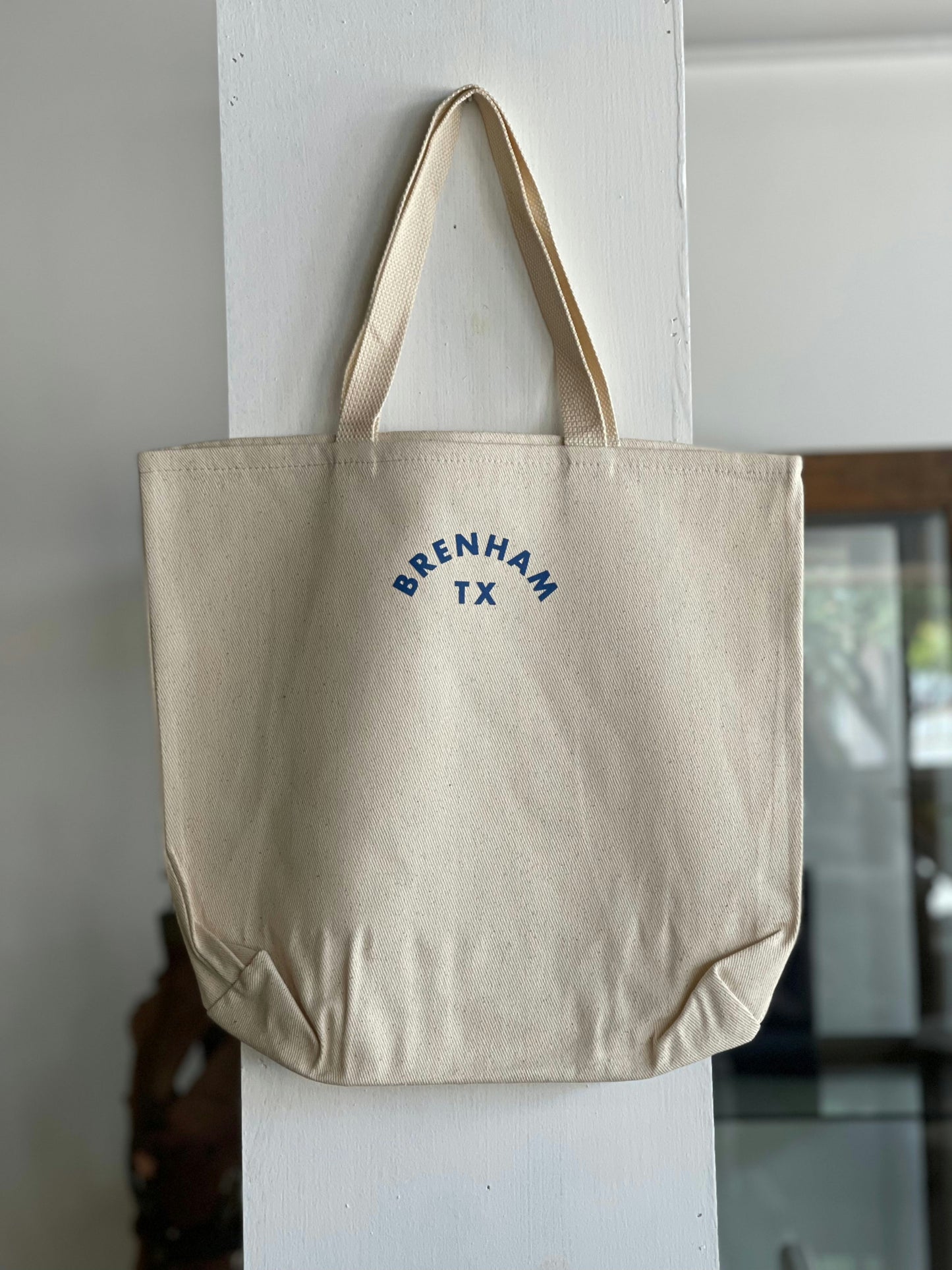 The Ballad of the Bird Dog Ballad Round Logo Tote, a beige cotton twill bag with blue "BRENHAM TX" print, hangs on a white wooden post indoors.