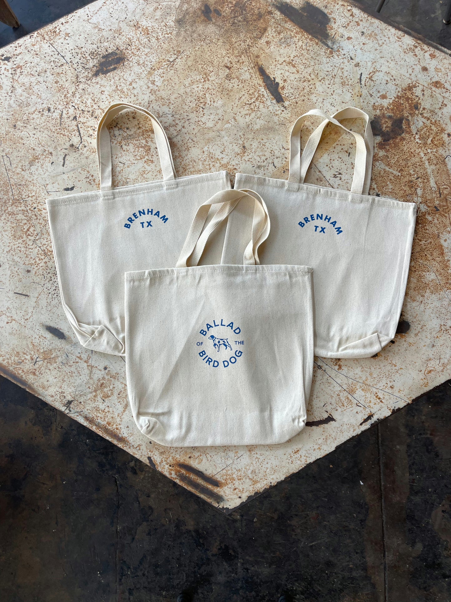Three beige cotton twill totes by Ballad of the Bird Dog rest on a wooden surface; two read "BRENHAM TX" and one has the Ballad Round Logo. Ideal for anyone seeking a stylish Brenham tote.