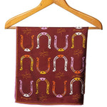 The Jenni Earle Bandana | Big Horseshoe features a maroon, hand-illustrated design with multicolored horseshoes and yellow swirls, finished with roulottage edges, shown hanging on a wooden hanger.