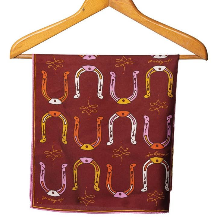 The Jenni Earle Bandana | Big Horseshoe features a maroon, hand-illustrated design with multicolored horseshoes and yellow swirls, finished with roulottage edges, shown hanging on a wooden hanger.