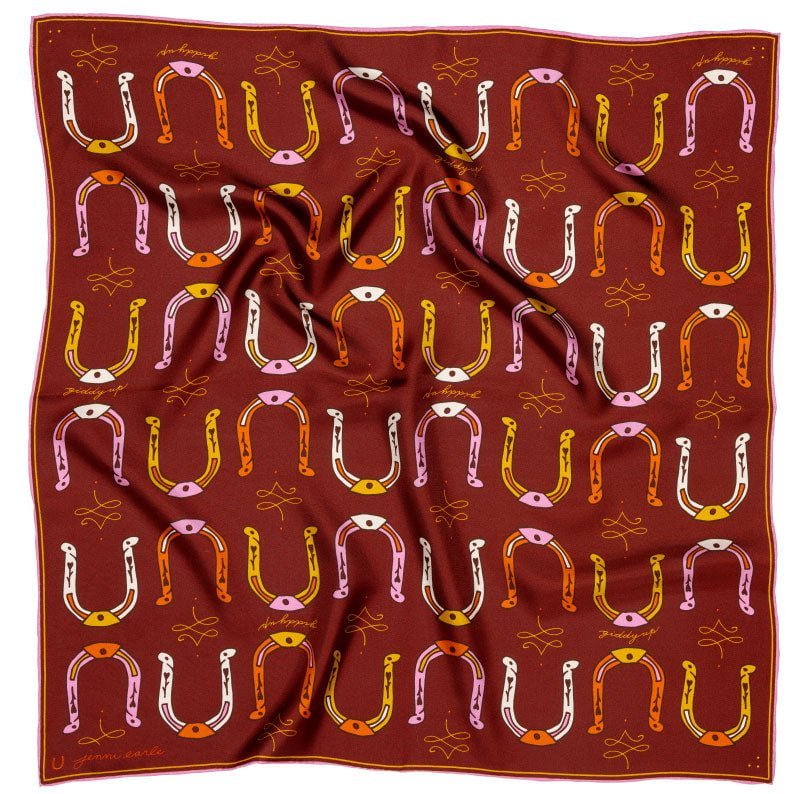 The Bandana | Big Horseshoe by Jenni Earle is a red silk scarf featuring roulottage edges and a repeating hand-illustrated pattern of yellow, pink, and white horseshoes, eyes, and lines. The scarf is slightly wrinkled.