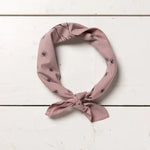 The Jenni Earle "Chin Up Buttercup" bandana, in light mauve cotton with tiny black sun prints, is knotted simply and displayed on a white wooden surface.