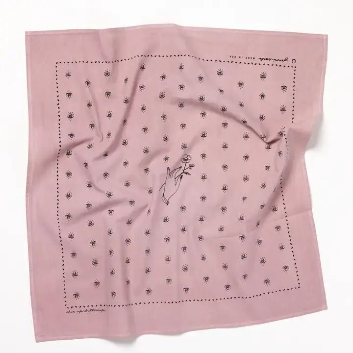 The "Chin Up Buttercup" bandana by Jenni Earle is a square pink cotton bandana with a central hand-holding-flowers line drawing, dotted border, and repeating floral pattern inspired by classic western styles.