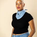 A person with short blonde hair wears a black short-sleeve top, blue jeans, and the ethically made "Fuck All Y'All" Bandana by Jenni Earle around their neck, standing against a beige background.