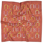 The "Giddy Up" bandana by Jenni Earle is a square scarf with a red background, featuring colorful horseshoes and abstract shapes—ideal for anyone who loves unique accessories or wants a thoughtful gift.