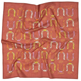 The "Giddy Up" bandana by Jenni Earle is a square scarf with a red background, featuring colorful horseshoes and abstract shapes—ideal for anyone who loves unique accessories or wants a thoughtful gift.